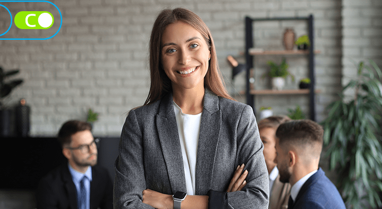 Na imagem, uma mulher vestida socialmente, com os cabelos castanhos claros, e os braços cruzados a frente da imagem, e ao fundo desfocado, há uma mesa de pessoas vestidas socialmente fazendo uma reunião.