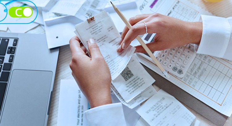 Na imagem, uma mesa, com v´rias notas fiscais em cima, e uma mão de mulher está analisando uma das notas com um lápis na mão também.